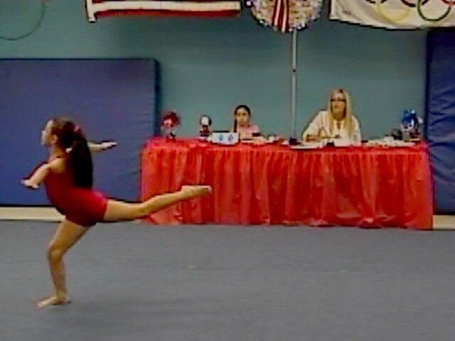 A Tumble Masters gymnast holding a scale on the floor in front of the judges during a Champion Day competition